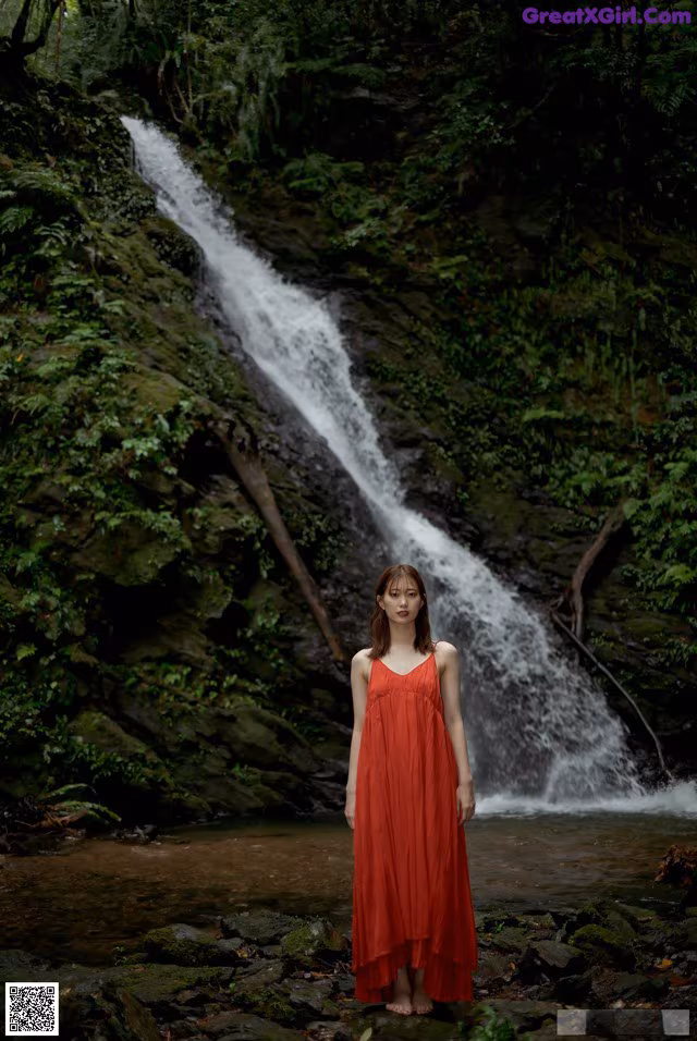 A woman in a red dress standing in front of a waterfall.